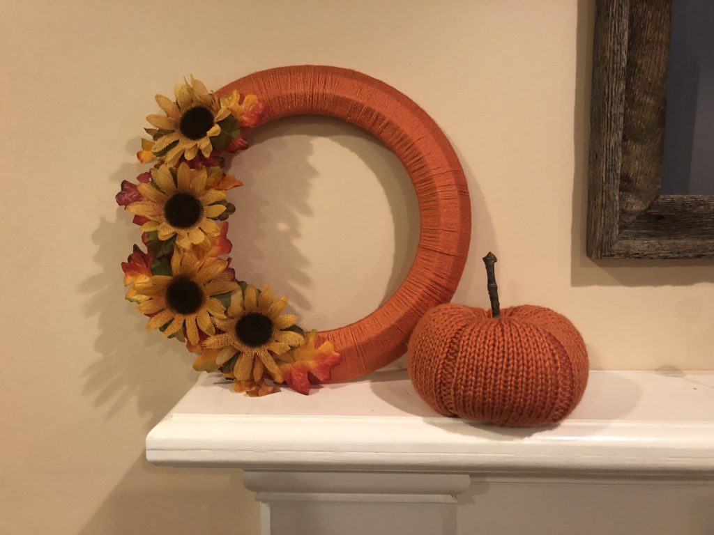 a yarn-wrapped wreath with fall leaves and sunflowers beside a knitted pumpkin on a mantel