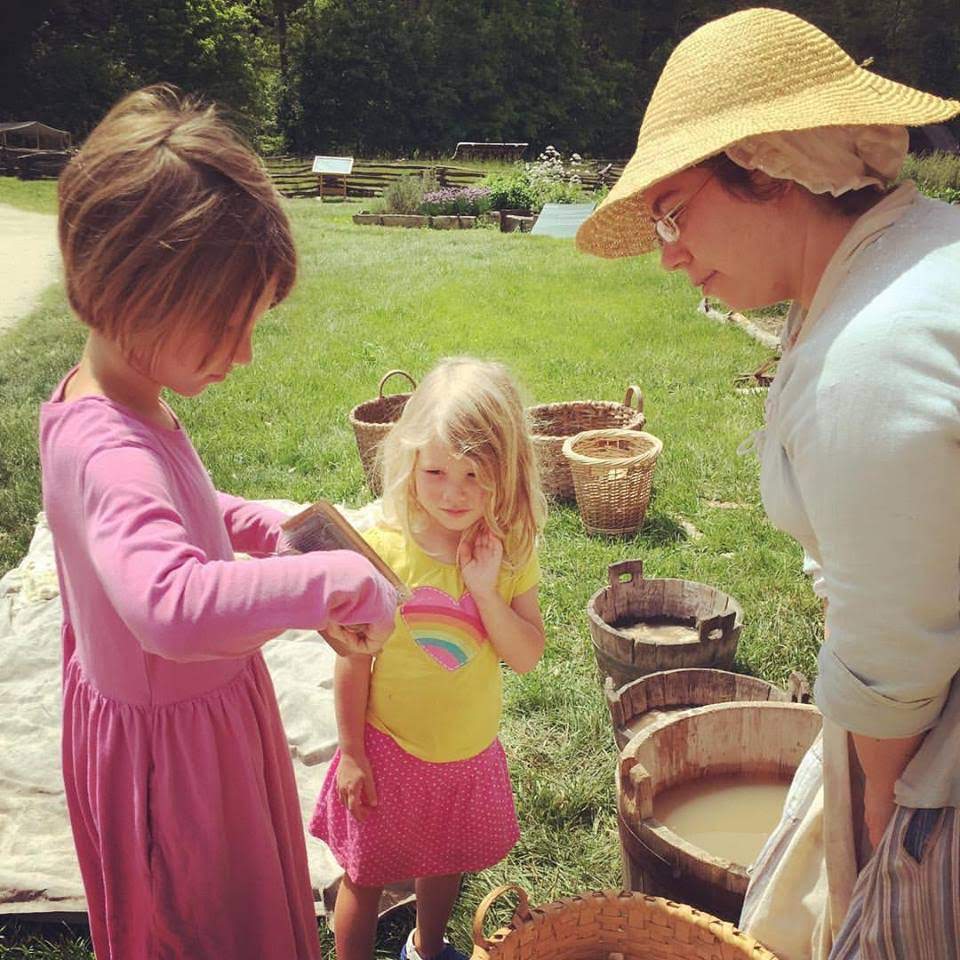 a child cards wool with hand carders while another child and a historical interpreter look on