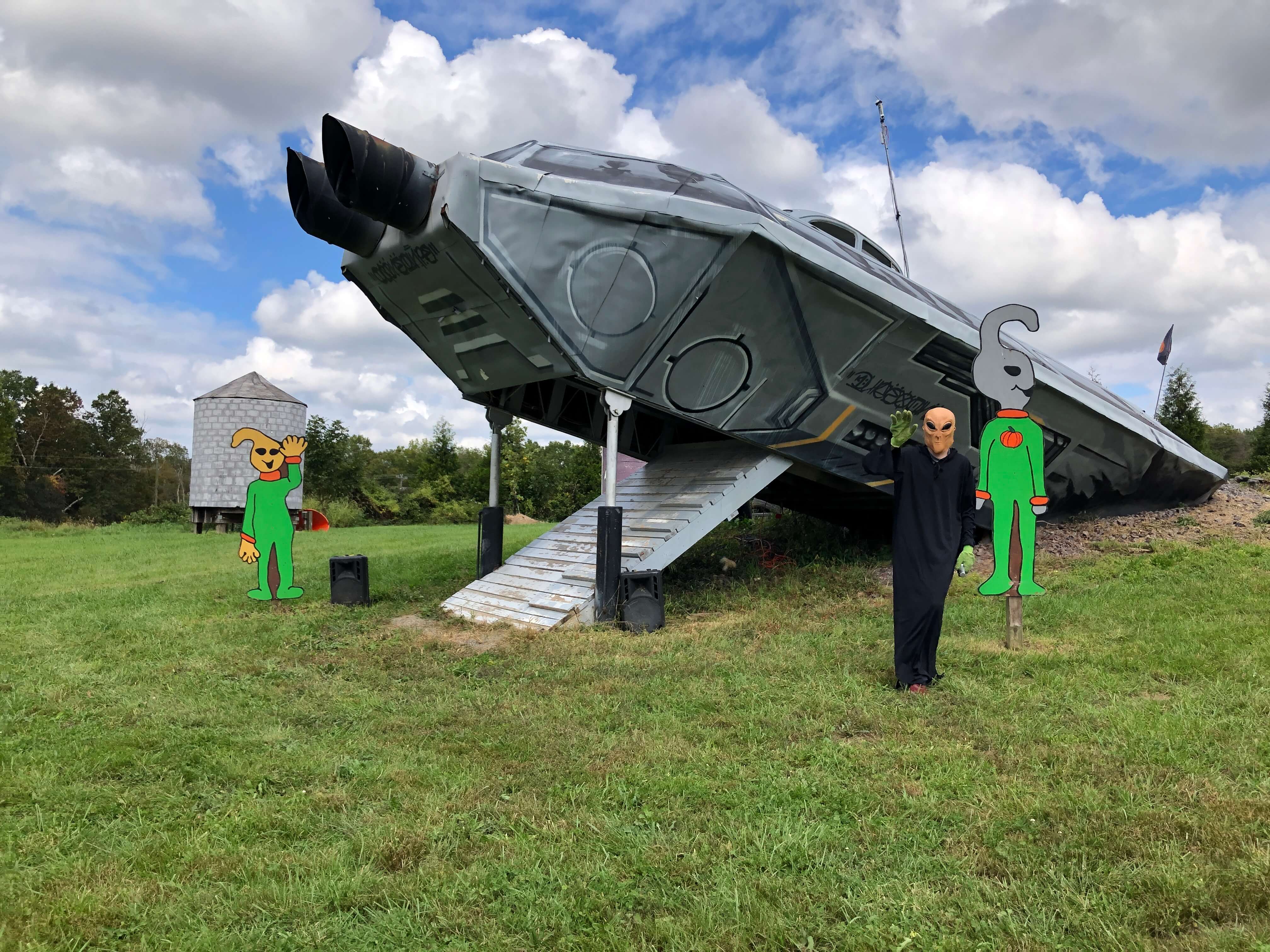 two alien cut-outs and one alien figure stand in front of a spaceship that appears to have crashed into the grassy hillside