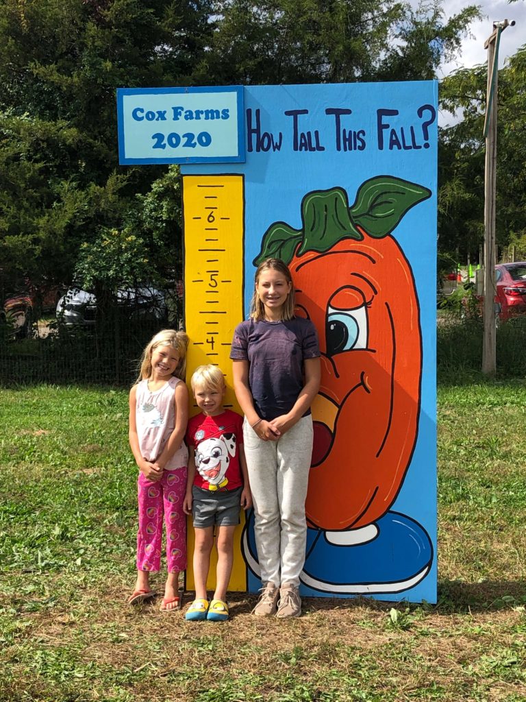 Three children pose in front of a sign with markings up to 6.5 feet. It reads "Cox Farms 2020 How Tall This Fall?"