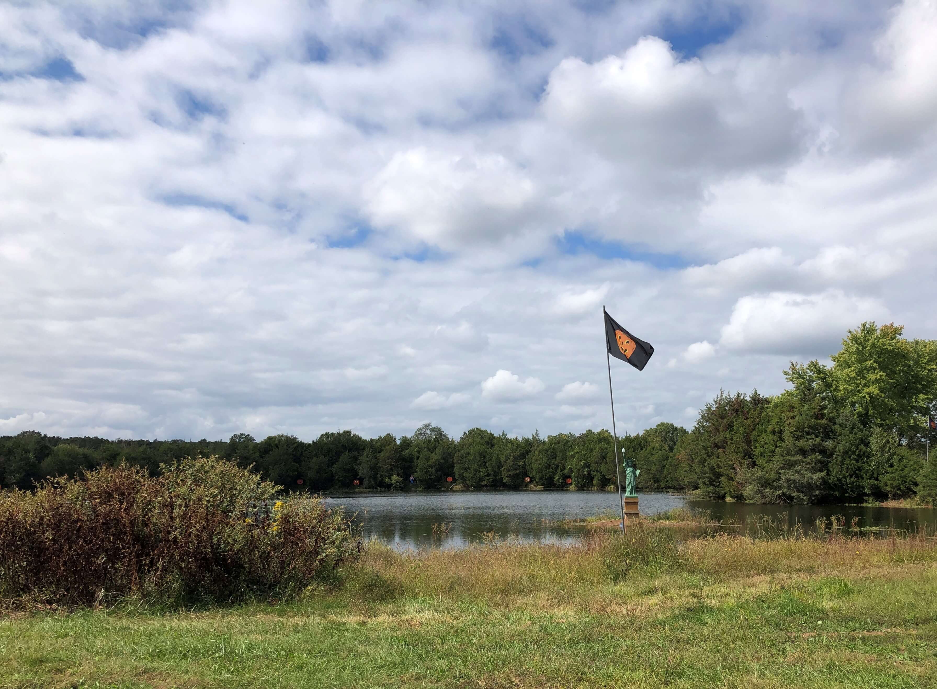 a small Statue of Liberty holds her torch high over a farm pond while a jack-o-lantern flag flies high