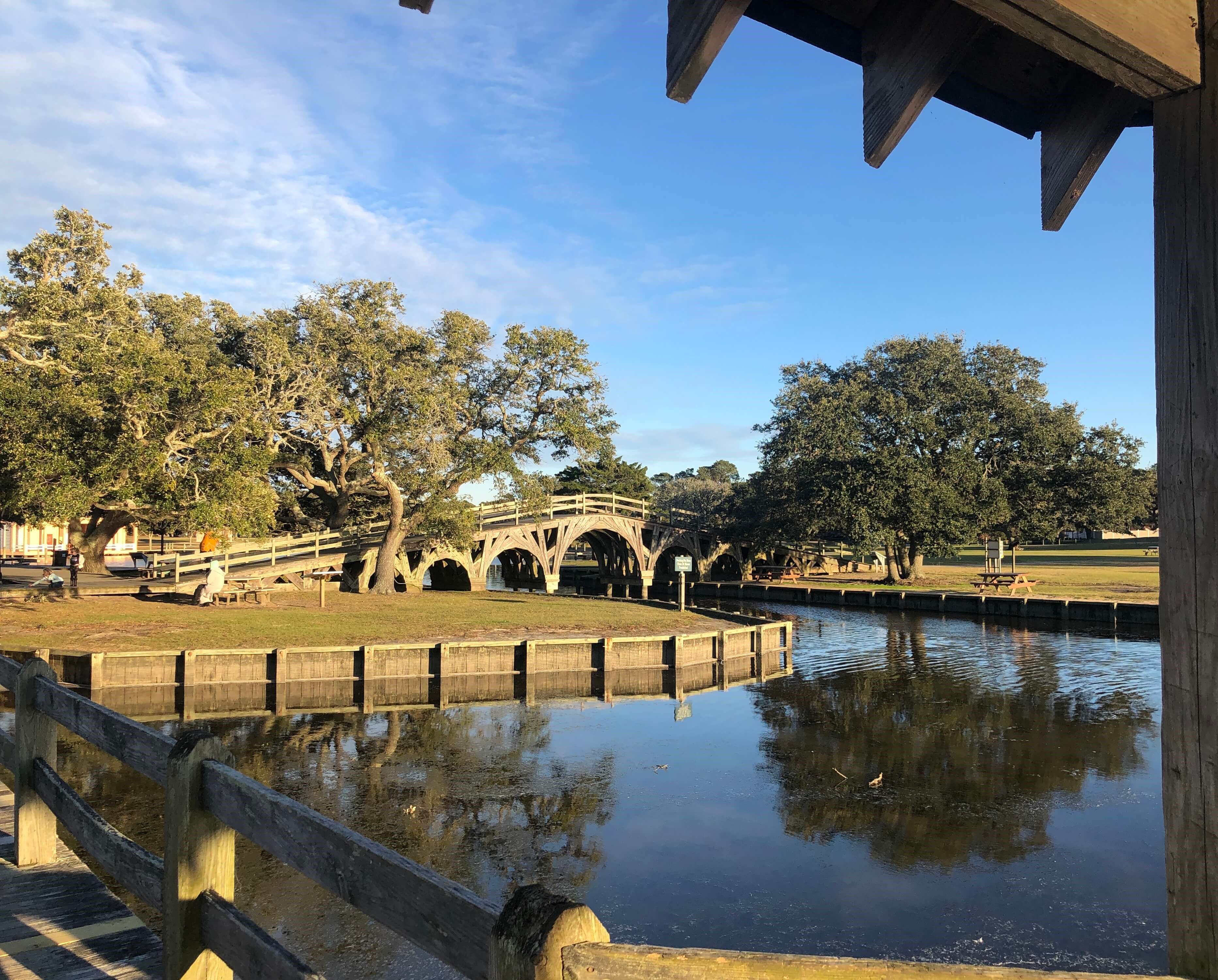 a wooden bridge lined with trees crosses an inlet while picnic tables surround